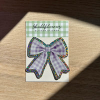 Glitter and gingham bow sticker on a checkered card with a wooden surface background