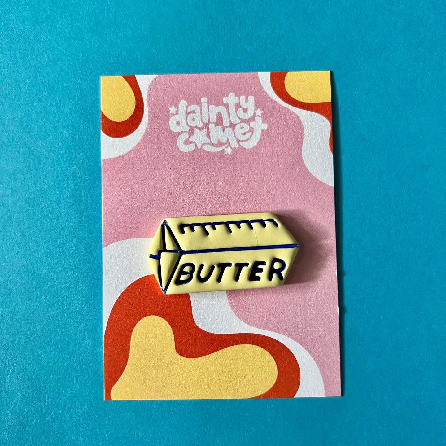 Pale yellow needle minder in the shape of a pat of butter with 'BUTTER' text, on a colorful card with 'Dainty Comet' branding against a blue background.