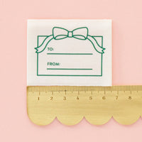 Gift tag label with a green bow design and 'TO' and 'FROM' fields, placed on a pink background with a gold ruler at the bottom.