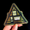 Holographic sticker of a haunted house with ghosts in a triangle shape held by a hand against a black background.