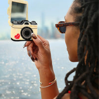 Person holding an open compact mirror in the shape of a vintage camera with a scenic background