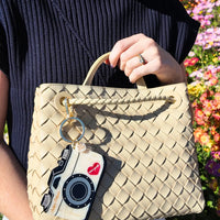 Beige woven handbag with camera-shaped keychain attached held by a person with a floral background