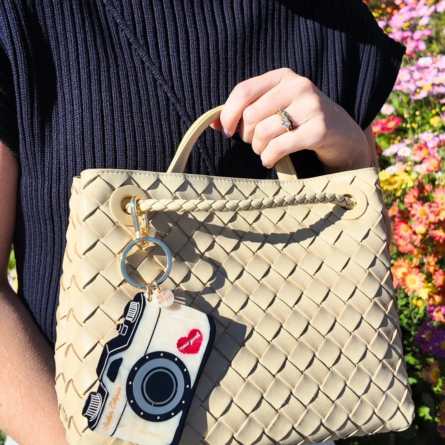 Beige woven handbag with camera-shaped keychain attached held by a person with a floral background
