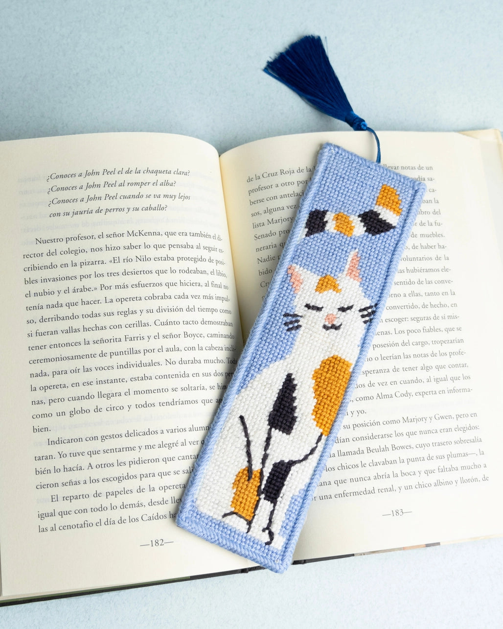 Needlepoint bookmark with a calico cat design on an open book with a light blue background