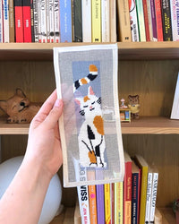 Hand holding a needlepoint cat design in front of a bookshelf