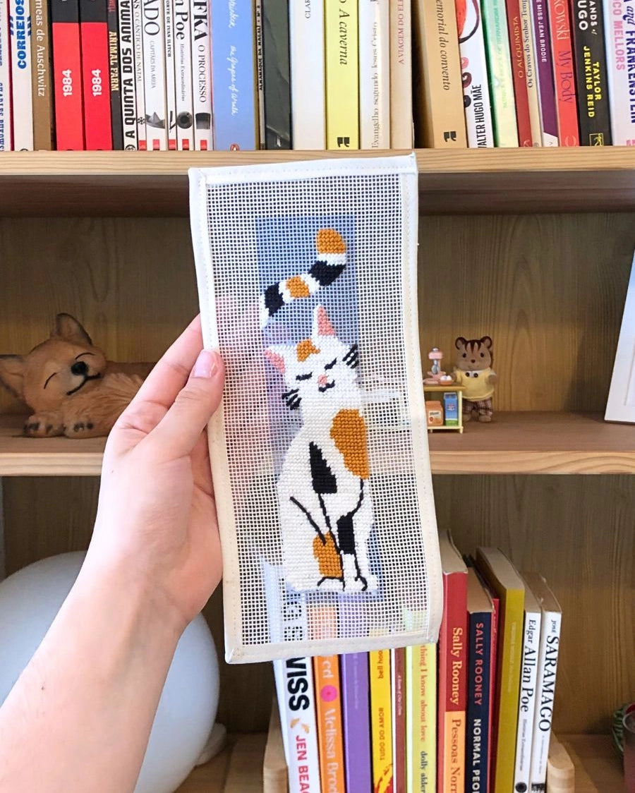 Hand holding a needlepoint cat design in front of a bookshelf