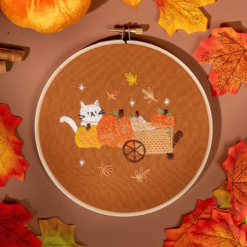 An embroidery hoop with a design featuring a white cat hiding behind large pumpkins, set against a warm brown cloth