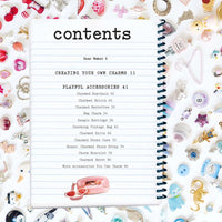 An open spiral-bound notebook showing a printed contents page surrounded by many pastel charms on a white background.