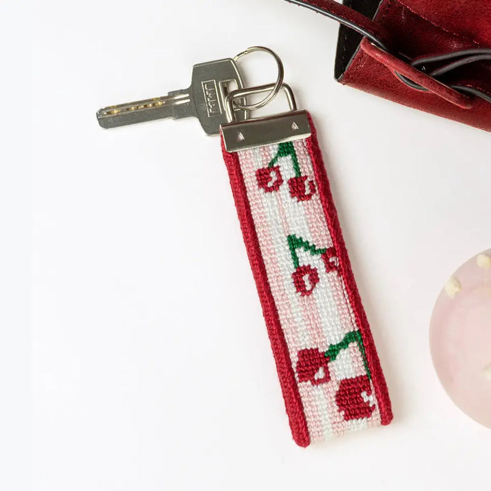 A cherry-patterned needlepoint key fob with a silver keyring and key, styled on a white surface with a red purse and a pink stone partially visible.