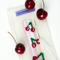 A close-up of a needlework kit showing a pink and white striped mesh canvas with a partially stitched cherry design in red, green, and white thread. A silver needle rests on the canvas, and three red cherries are placed around it.