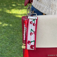 A close-up of a key fob strap on a handbag featuring a cherry-themed needlepoint design on a red leather bag, next to another charm with pink beads, and a small gold cherry charm, worn outdoors with blue jeans and a cream knit sweater.