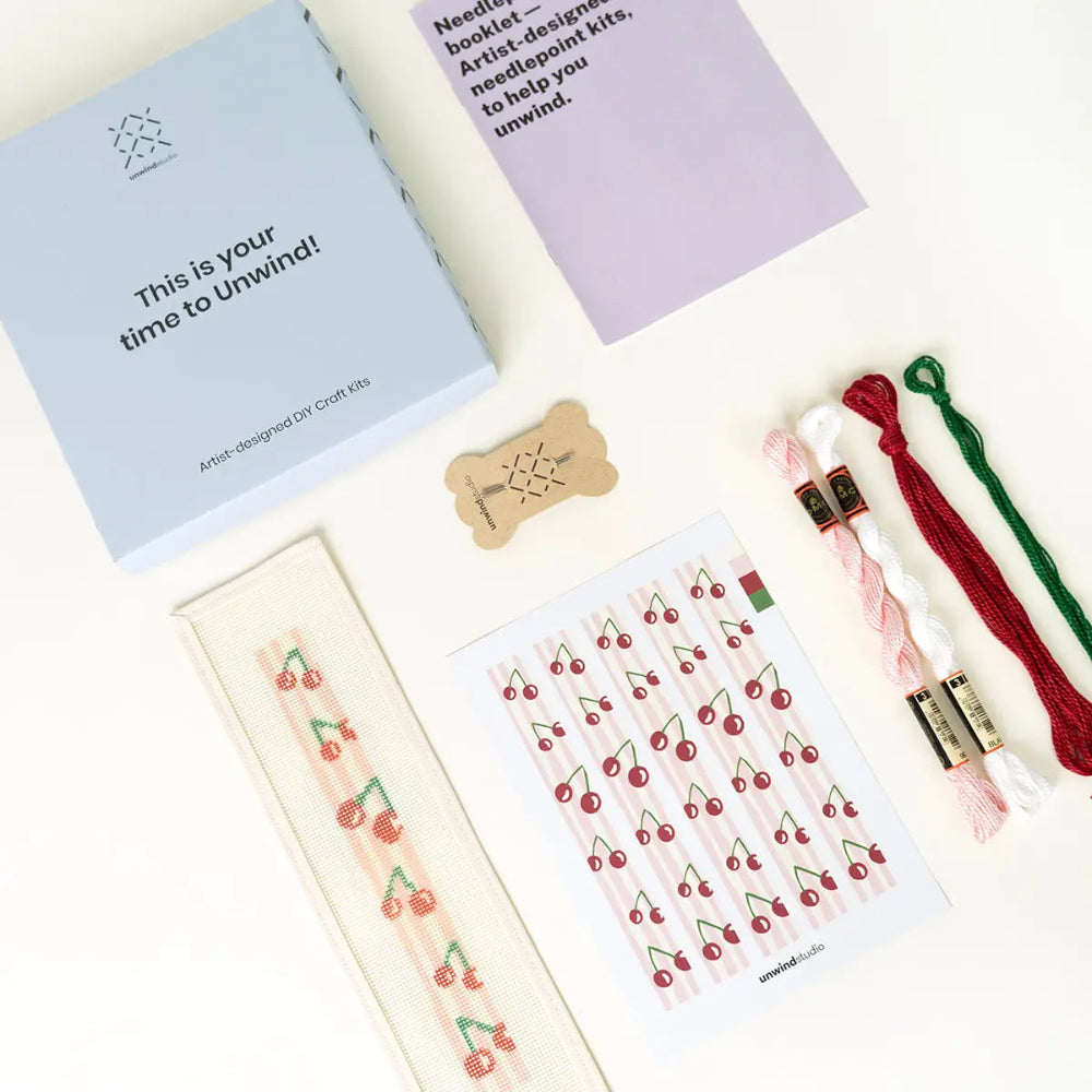 A flat-lay view of a needlepoint design kit showing its components on a white background: a light blue box with the text 'This is your time to Unwind!', a lavender needlepoint booklet, a cherry-patterned canvas, a sheet of cherry stickers, and three spools of embroidery thread in white, pink, and green.