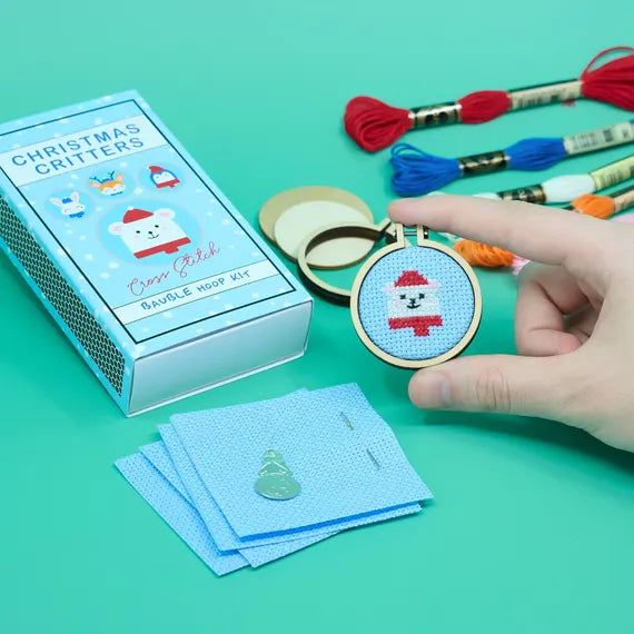 A blue box with the words "Christmas Critters" and illustrations of animals on it, a few skeins of embroidery thread, a few squares of fabric, and a hand holding an embroidery hoop with a cross stitch polar bear design on an aqua background.