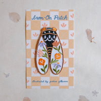 An embroidered iron-on patch shaped like a Cicada, displayed on its printed backing card. The patch features orange and purple flowers, green leaves, and a black and gold designed center.