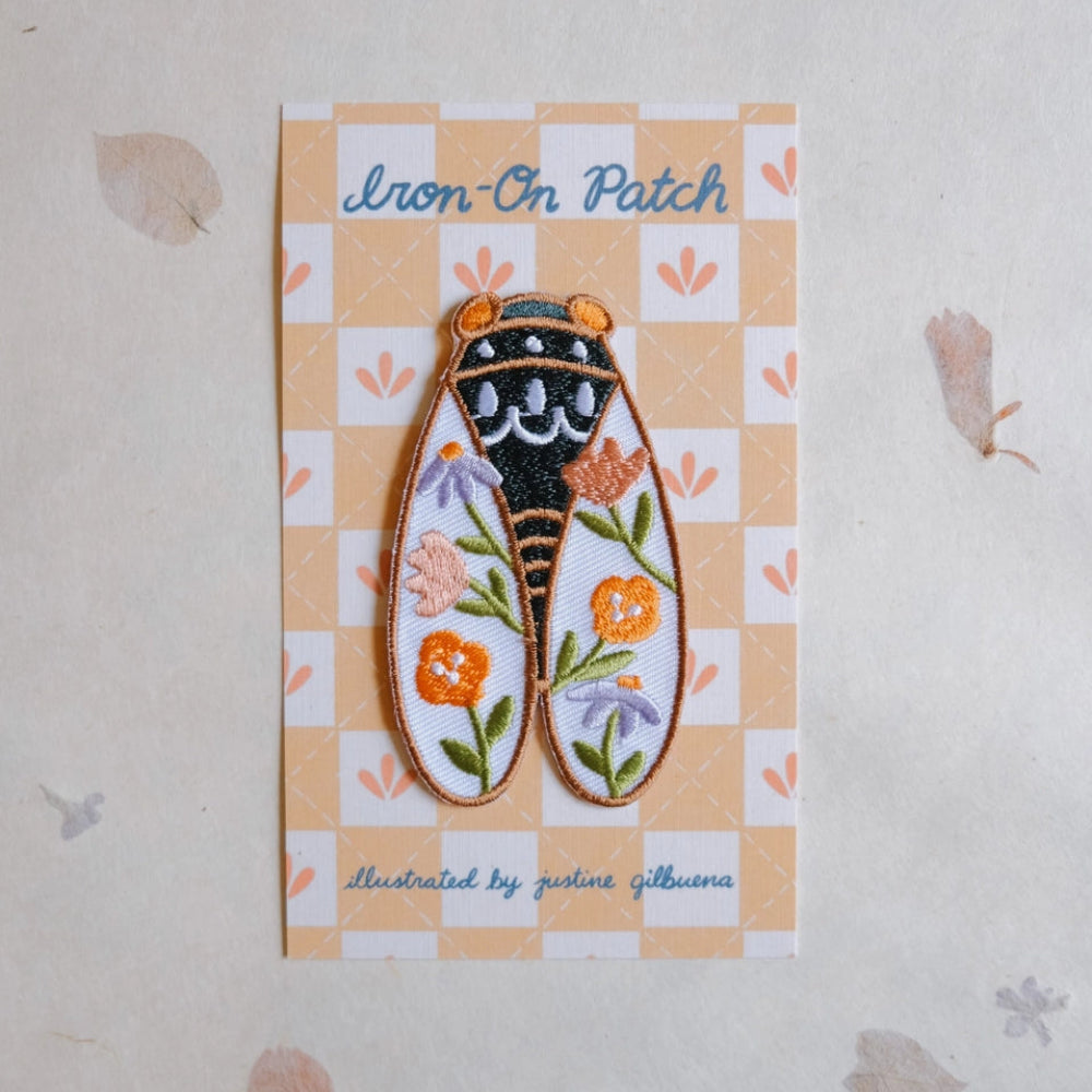 An embroidered iron-on patch shaped like a Cicada, displayed on its printed backing card. The patch features orange and purple flowers, green leaves, and a black and gold designed center.