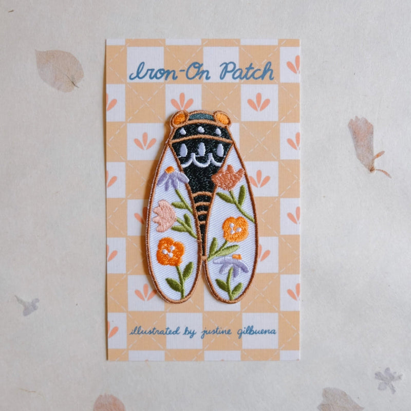 An embroidered iron-on patch shaped like a Cicada, displayed on its printed backing card. The patch features orange and purple flowers, green leaves, and a black and gold designed center.