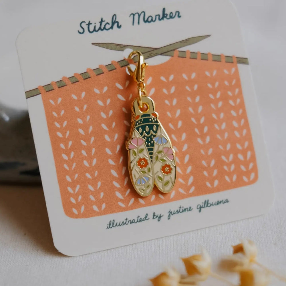 A gold-tone enamel stitch marker charm shaped like a cicada with floral details, mounted on a white display card labeled 'Stitch Marker' with an orange knitted pattern illustration.