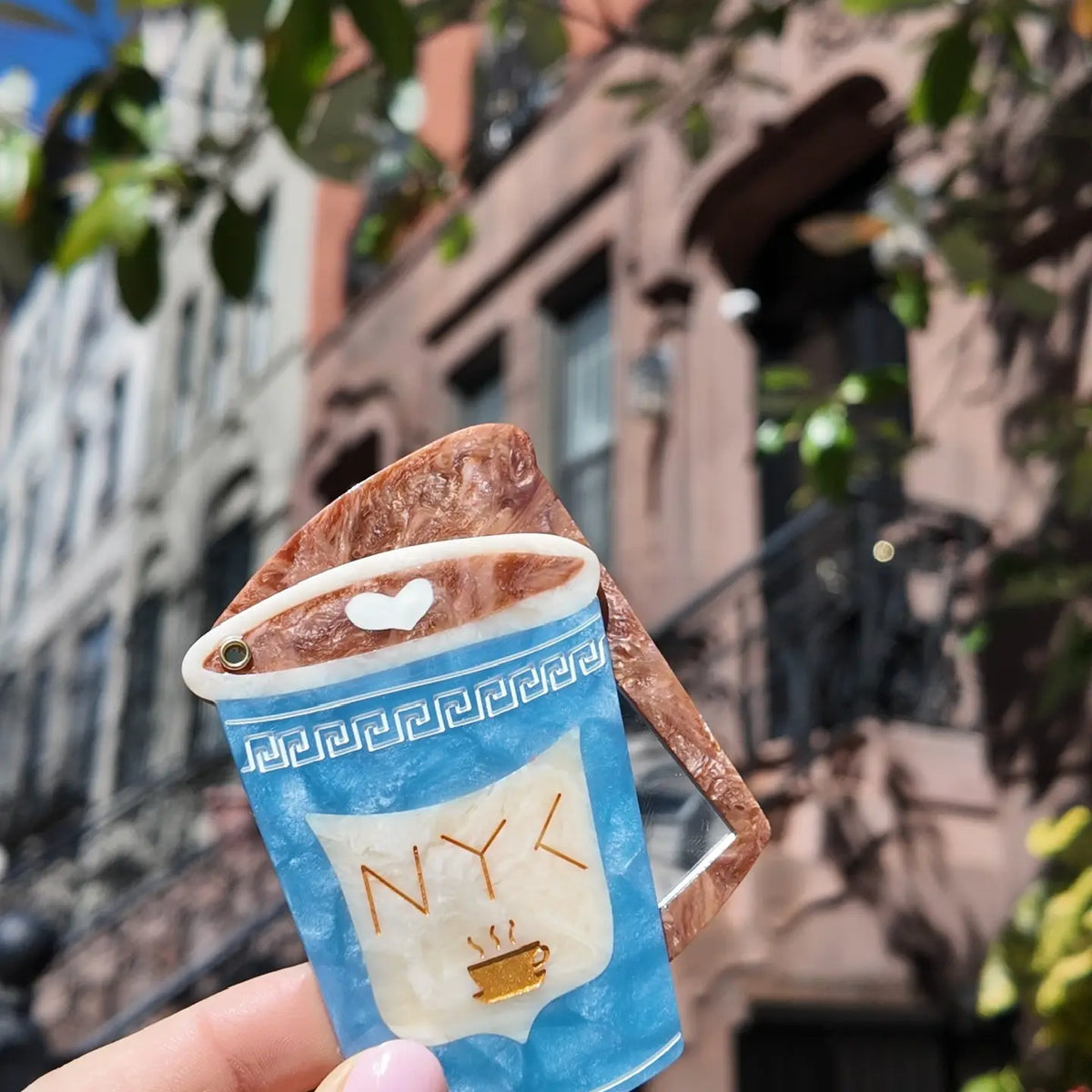 Blue and white compact mirror shaped like a coffee cup with text reading "NYC" in gold, held in front of a blurred cityscape.