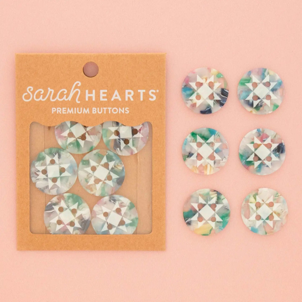 Sarah Hearts premium buttons displayed next to their kraft paper packaging on a solid peach-pink background. The buttons feature a colorful, marbled pattern with shades of green, blue, purple, and white, and are arranged in two groups: six inside the clear window of the brown cardboard packaging and six loose buttons to the right.
