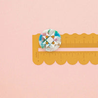 A round button with a geometric star pattern in white with marbled shades of blue, green, pink, and white, shown next to a yellow scalloped edge ruler on a solid pink background.