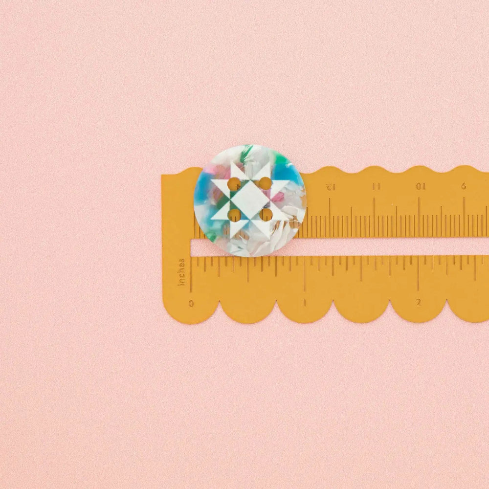 A round button with a geometric star pattern in white with marbled shades of blue, green, pink, and white, shown next to a yellow scalloped edge ruler on a solid pink background.