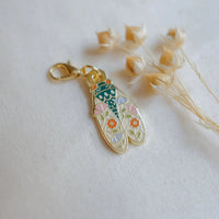 A gold-toned enamel charm shaped like a cicada decorated with pink, orange, green, and green floral details, resting on a white fabric next to dried beige flower stems.