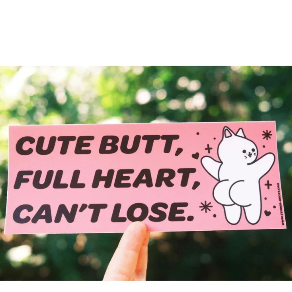 Pink sticker with text 'Cute butt, full heart, can't lose.' held against a blurred green background