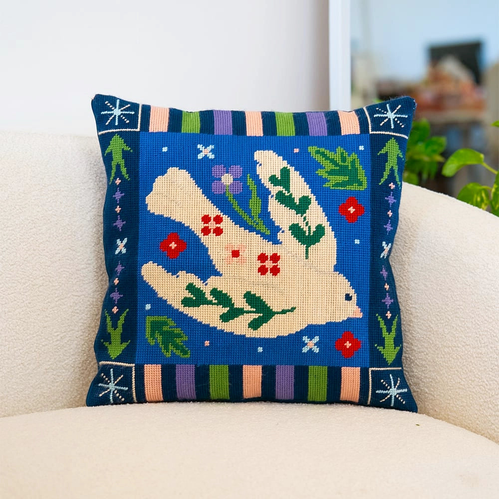 A needlepoint cushion with a blue floral design featuring a white dove in the middle of a blue background.