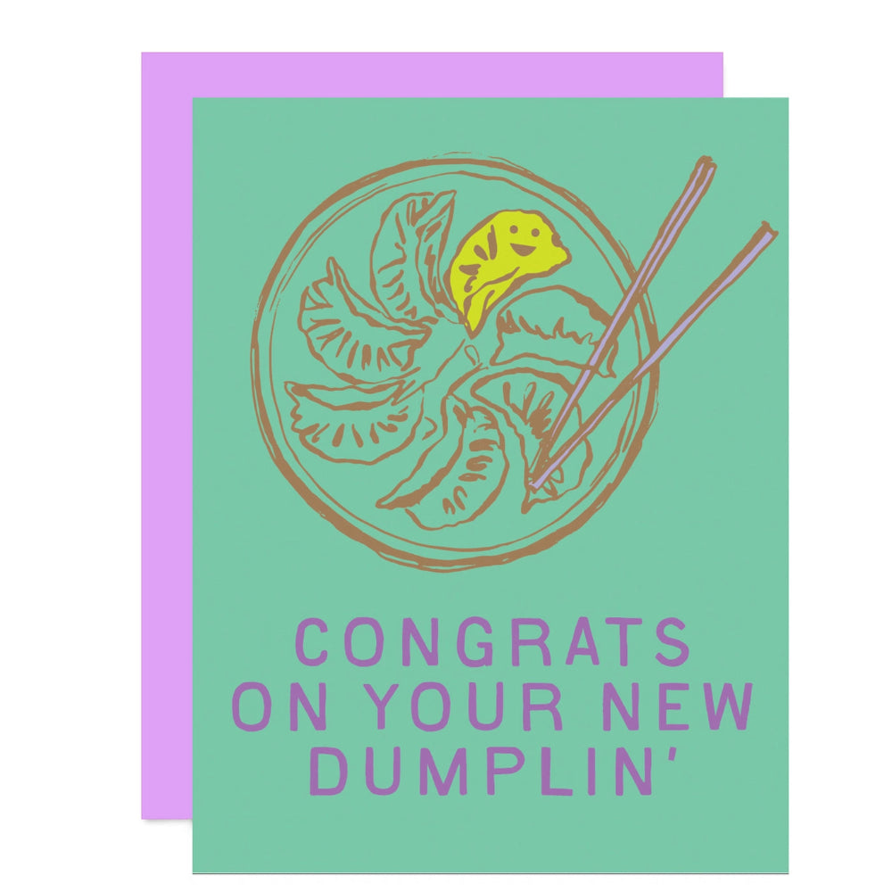 A greeting card with a light mint green front and a lavender envelope behind it. The card features a hand-drawn illustration of dumplings in a bowl with chopsticks and the text 'CONGRATS ON YOUR NEW DUMBLIN'' in lavender.