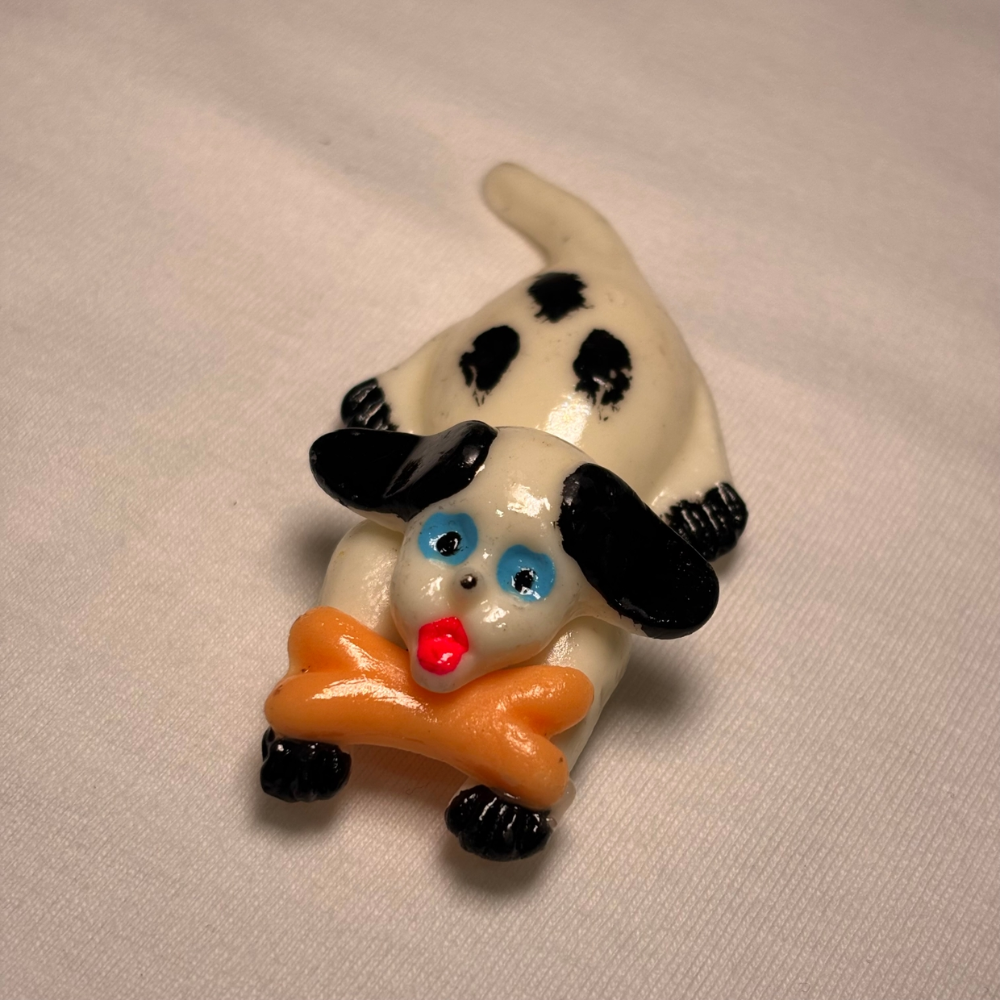 Small clay magnet of a white dog laying down holding a brown bone