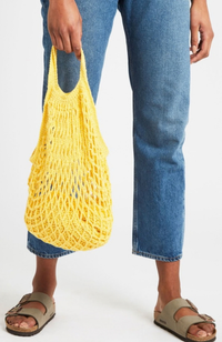 Model in jeans and sandals holding a bright yellow crocheted market bag on a white background.