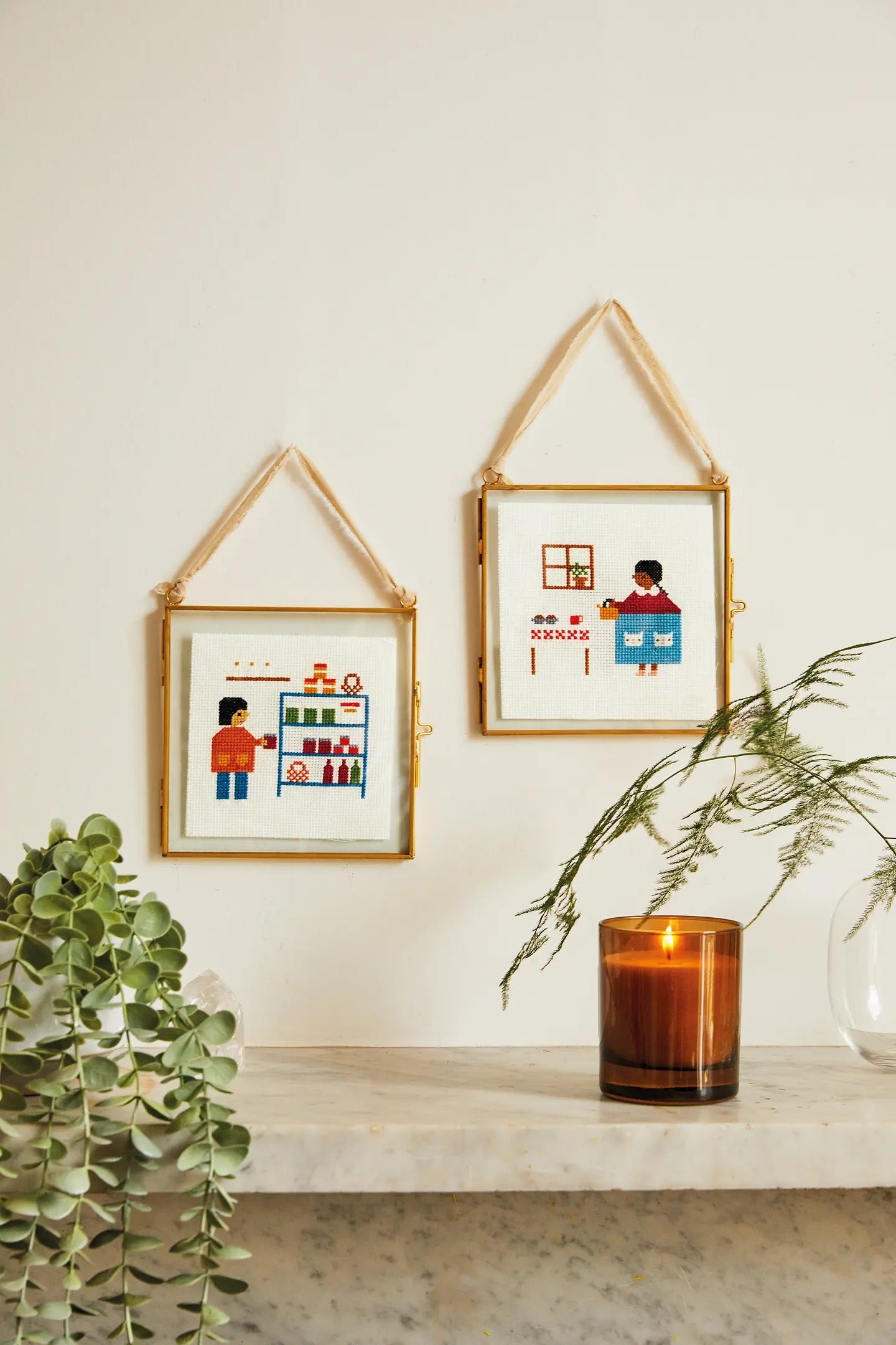 Two framed illustrations on a wall with a candle and plants in the foreground.