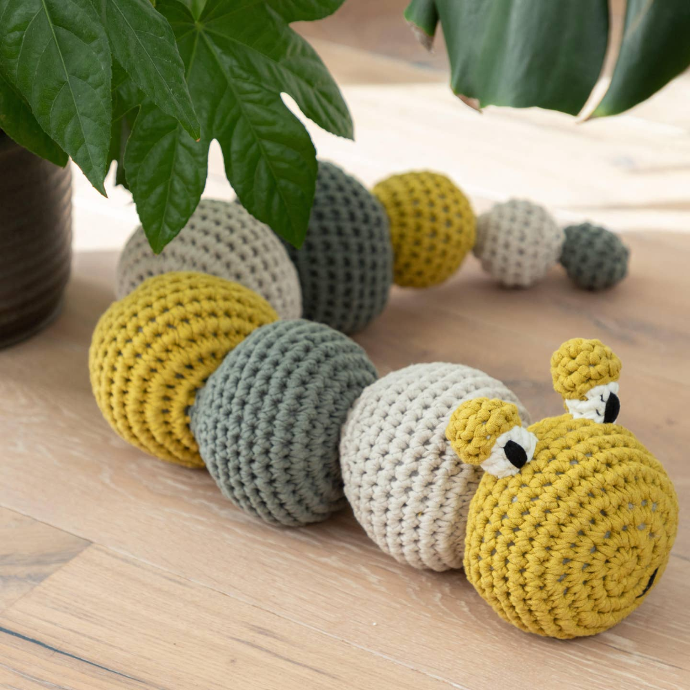 Yellow, grey, and green crochet caterpillar on wood floor with plant in background