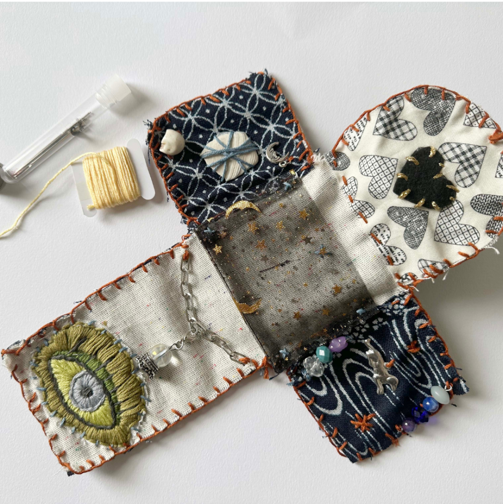 A handmade fabric jewelry organizer or pouch laid flat on a white surface, shown open to reveal multiple patterned fabric compartments with beads, thread, and small decorative elements. A spool of yellow thread and a small tool are placed beside it.