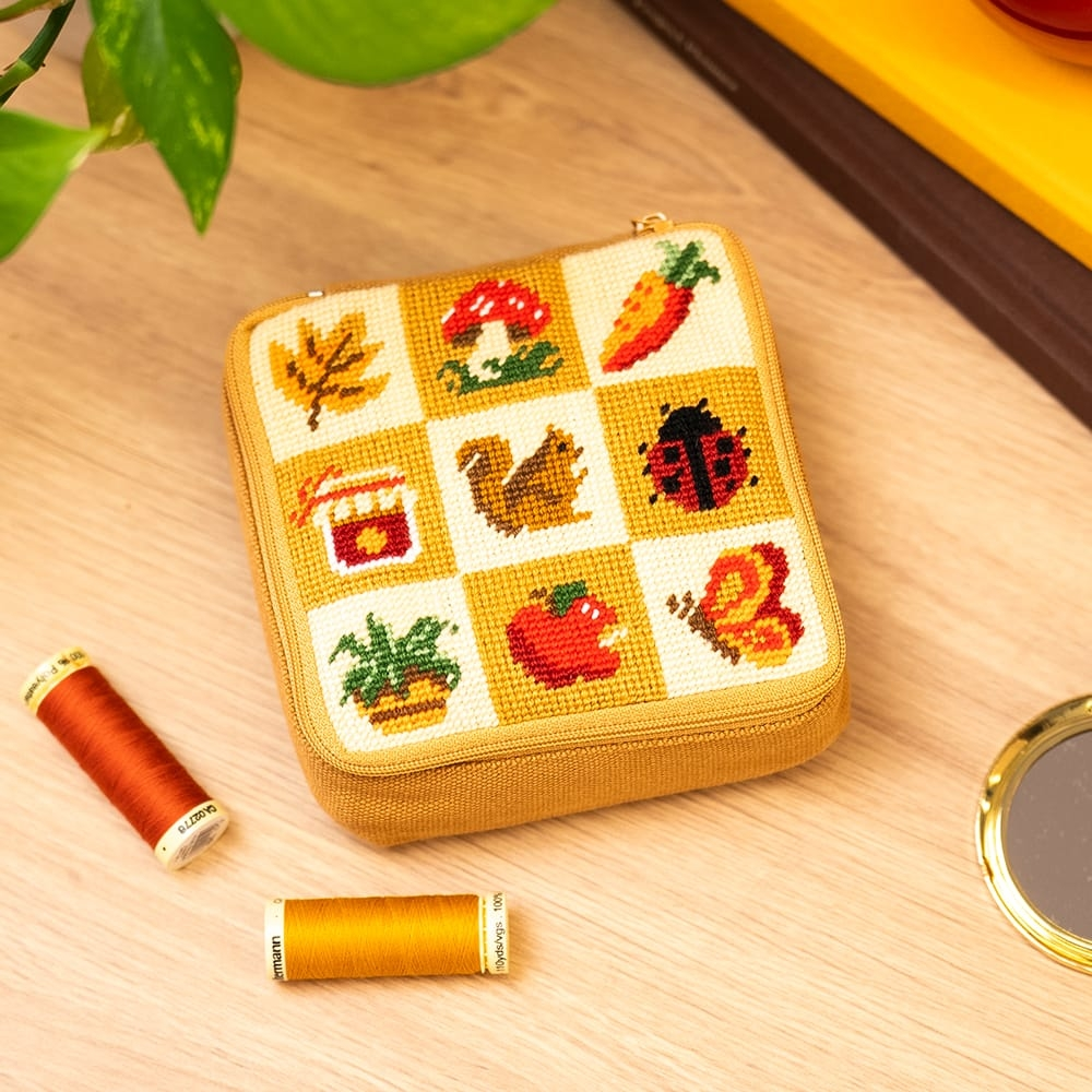 A square zipper pouch decorated with nine fall-themed images in needlepoint, including a squirrel, carrot, mushroom, and leaf