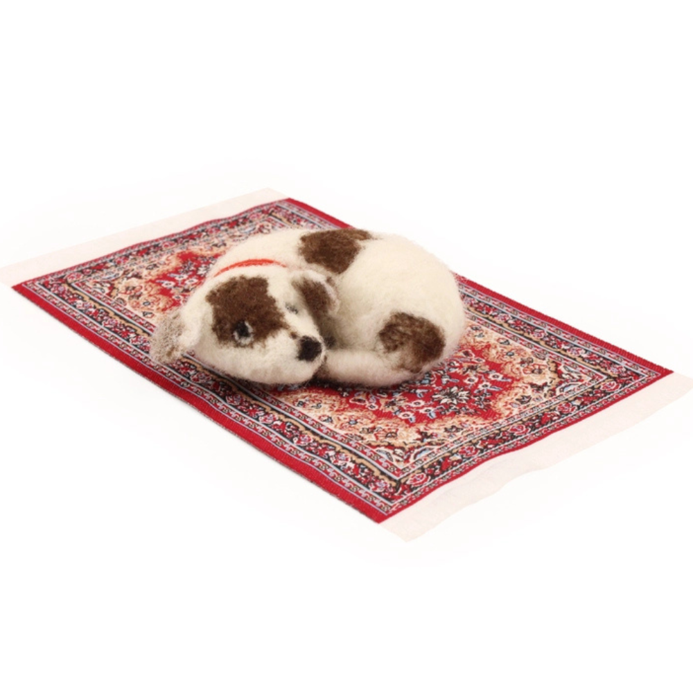A small brown and white felted puppy toy wearing a red collar, curled up asleep on a miniature red patterned rug against a plain white background.