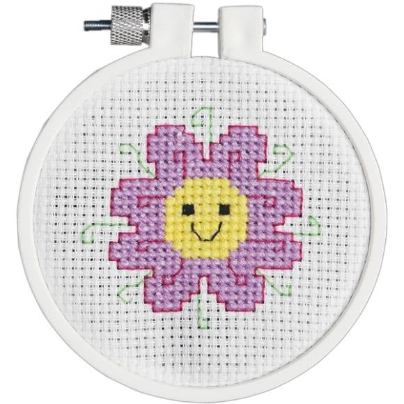 Flower cross-stitch design with yellow smiley face, 8 purplish petals with pink outline and light green tendrils.