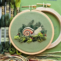Embroidery hoop with embroidered snail design on green fabric, surrounded by embroidery threads.