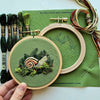 Embroidery hoop with green fabric featuring a nature-themed design, surrounded by embroidery threads and tools on a green mat.