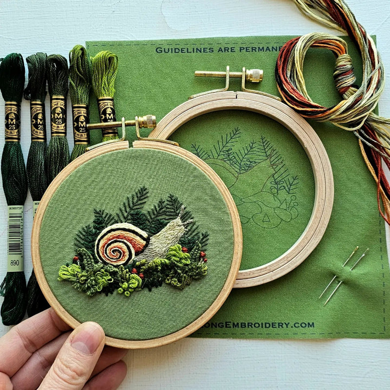 Embroidery hoop with green fabric featuring a nature-themed design, surrounded by embroidery threads and tools on a green mat.