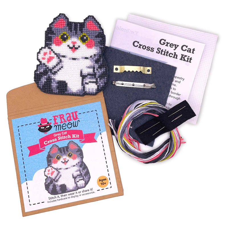 A grey cat cross stitch kit laid out on a white background, including a finished grey cat patch, printed kit cards, thread, pins, and a black fabric backing.