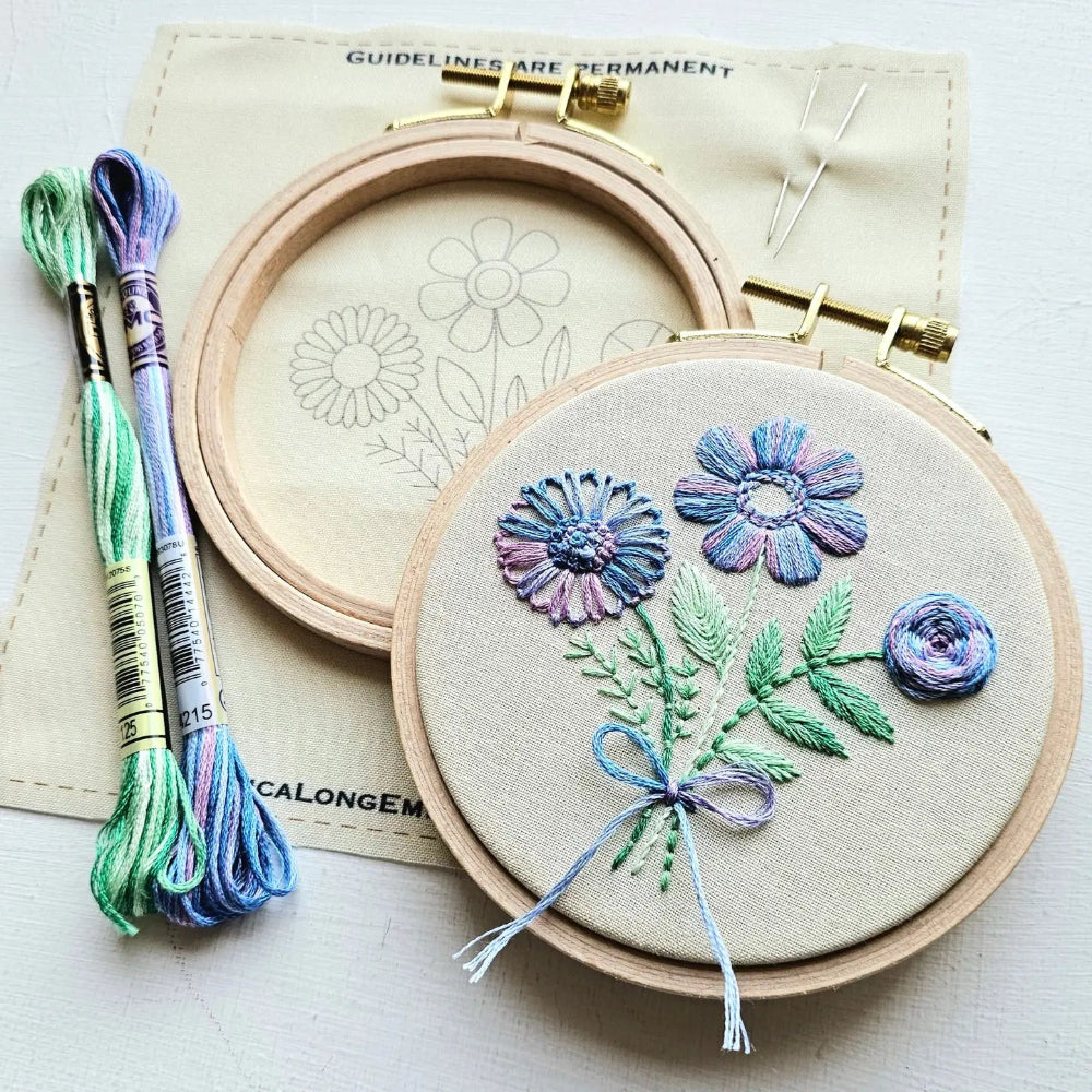 A close-up of a floral embroidery project in a wooden hoop, with two spools of variegated embroidery floss, a second empty hoop, and a printed pattern sheet on a light surface.
