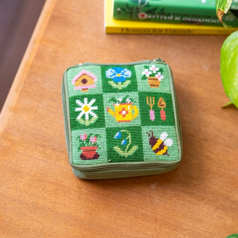 A square zipper pouch featuring a needlepoint design with a watering can, bumblebee, birdhouse, and flower pot among other images