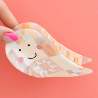 A translucent ghost hair claw clip with a hand-drawn smiling face, held by a hand with pink fingernails against a solid coral-pink background.