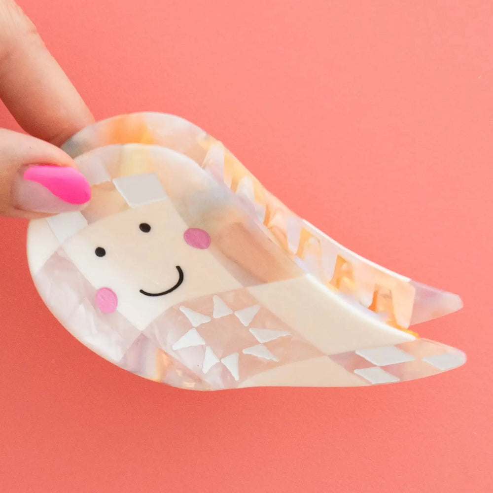 A translucent ghost hair claw clip with a hand-drawn smiling face, held by a hand with pink fingernails against a solid coral-pink background.