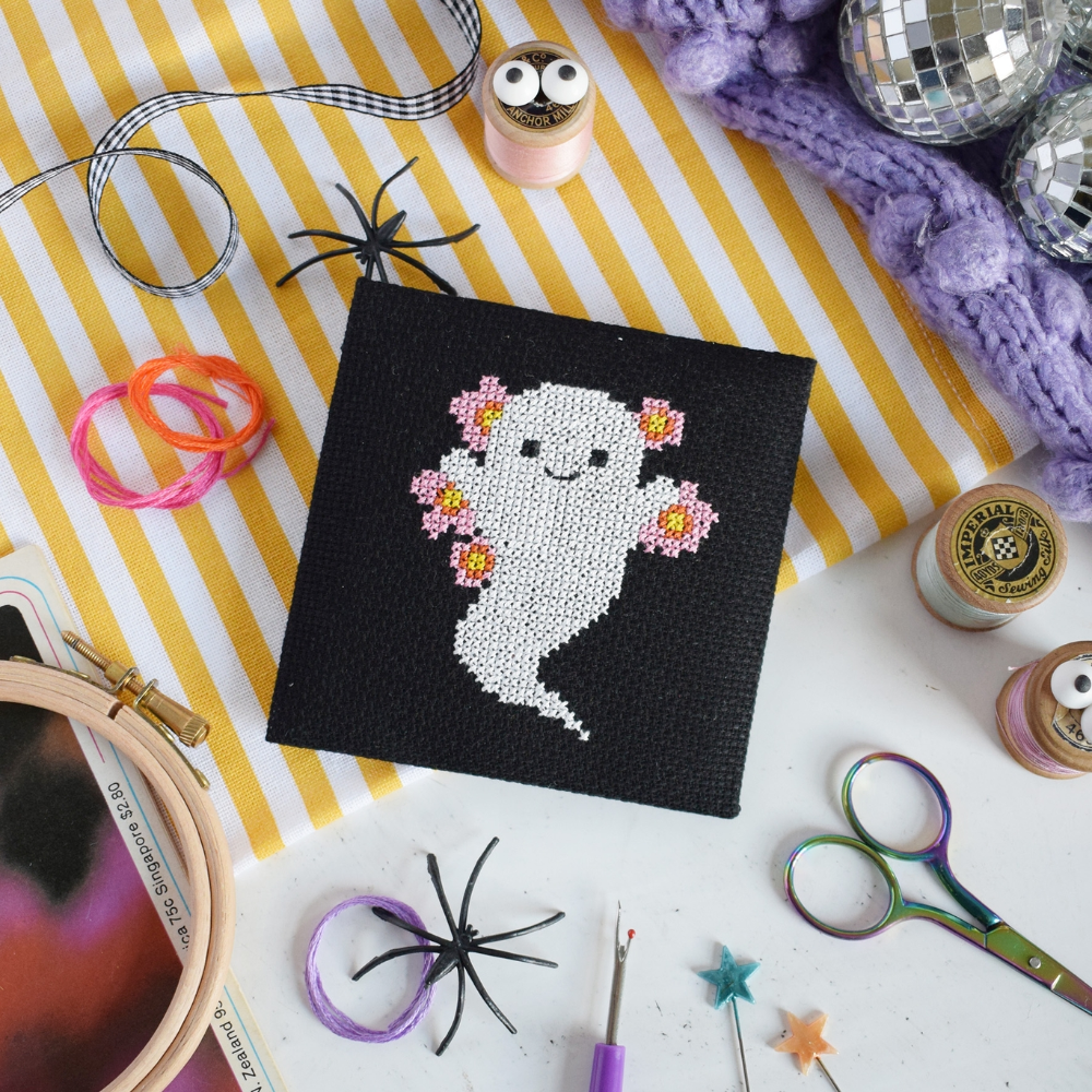 A square black cross-stitch canvas featuring a white ghost design surrounded by pink flowers, and by craft tools like scissors, pins, and thread in background.