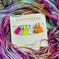 Six colorful ghost stitch markers sit against a background of brightly colored yarn