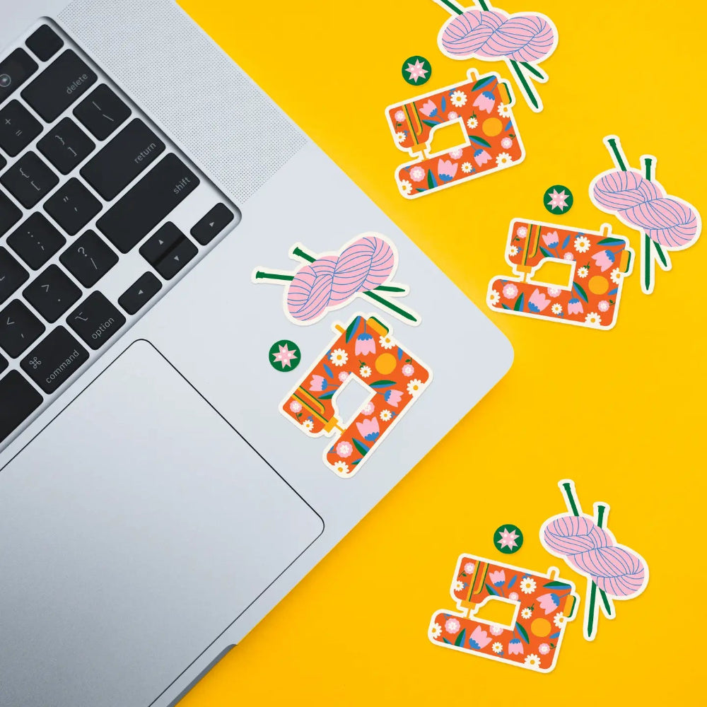 A silver laptop on a bright yellow background with decorative stickers showing a floral sewing machine and pink yarn hanks with knitting needles. The laptop keyboard is partially visible, and the stickers are arranged diagonally across the frame.