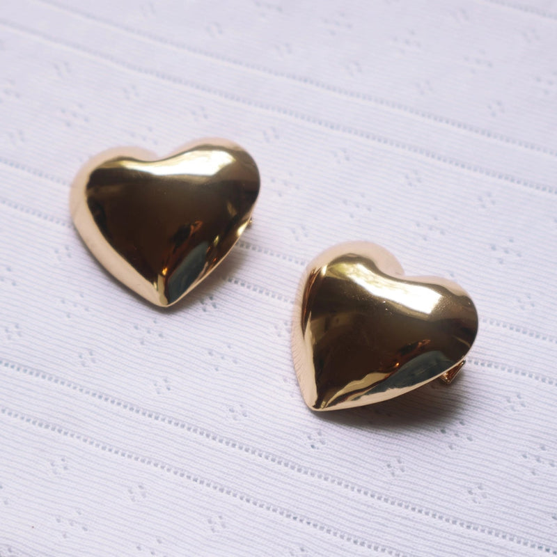 A pair of shiny gold heart-shaped stud earrings resting on a textured white fabric background.