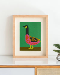 Framed needlepoint artwork with a colorful goose design resting on a white table against a white background